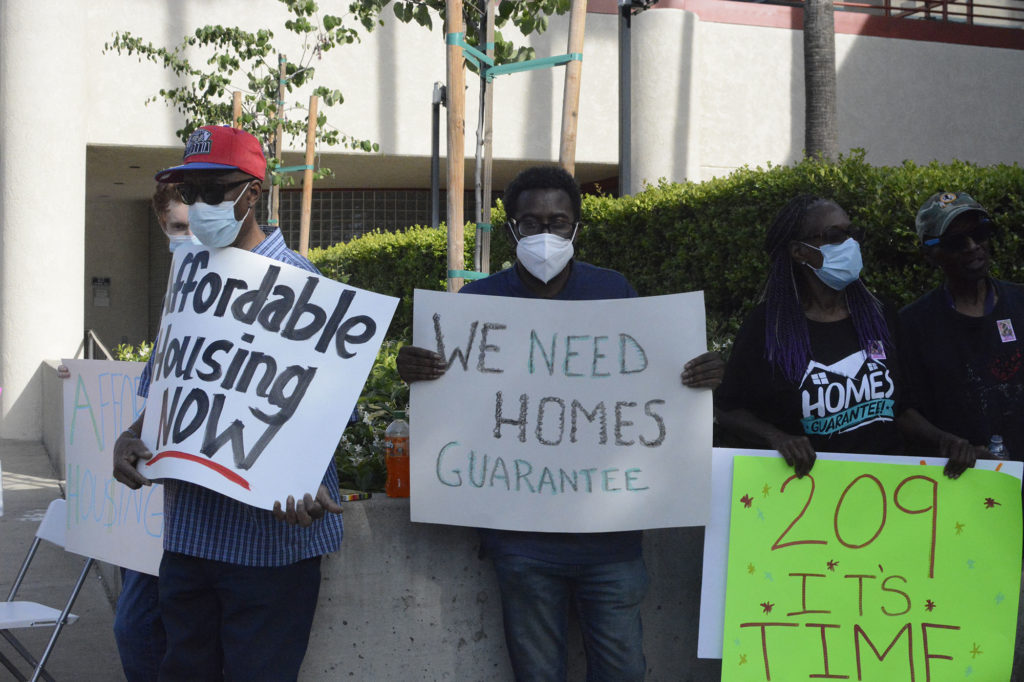 Three community members stand in front of Merced Civic center holding up signs that say: "affordable housing now," "WE NEED HOMES GUARANTEE," and "209 IT'S TIME"