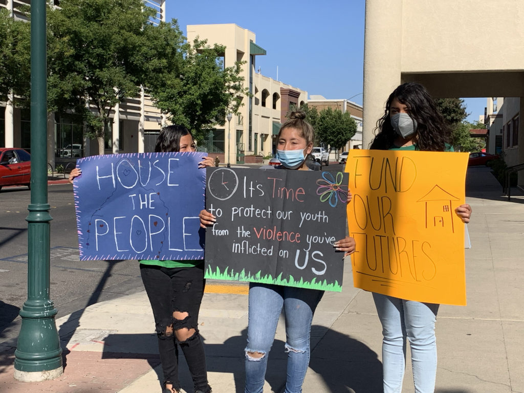 Three people are holding up the following signs: "House the People," "It's time to protect our youth from the violence you've inflicted on us," and "fund our futures."