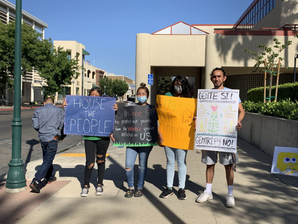 Four people are holding up the following signs: "House the People," "It's time to protect our youth from the violence you've inflicted on us," "fund our futures," "gente si! gentrífy no!"