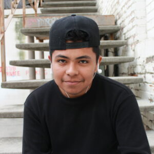 A young person named Carlos posing for his personal narrative piece in the stairs with his black hat and earphones on 